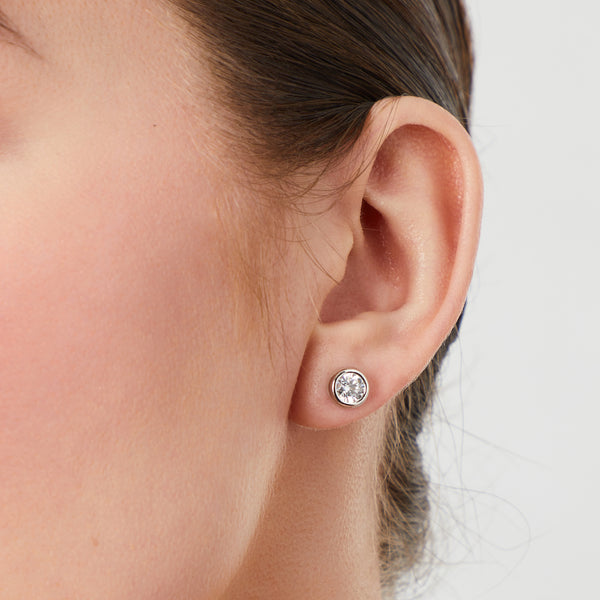 cloase up of model wearing a pair of 1 carat solitaire cubic zirconia stud earrings in a bezel setting on sterling silver.